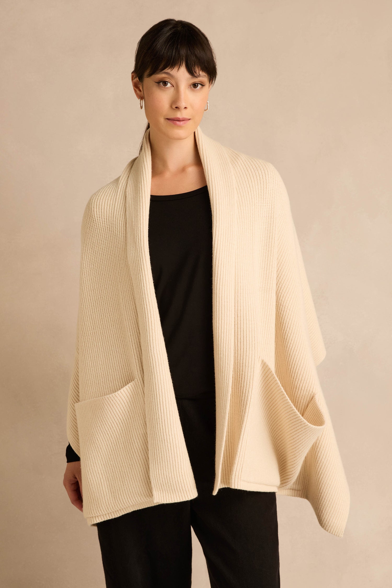 A woman stands against a plain beige background, wearing the Cream Pocket Scarf—an oversized pocket scarf in a cream-colored, ribbed knit made from a recycled fibre blend—draped over her black top and pants.