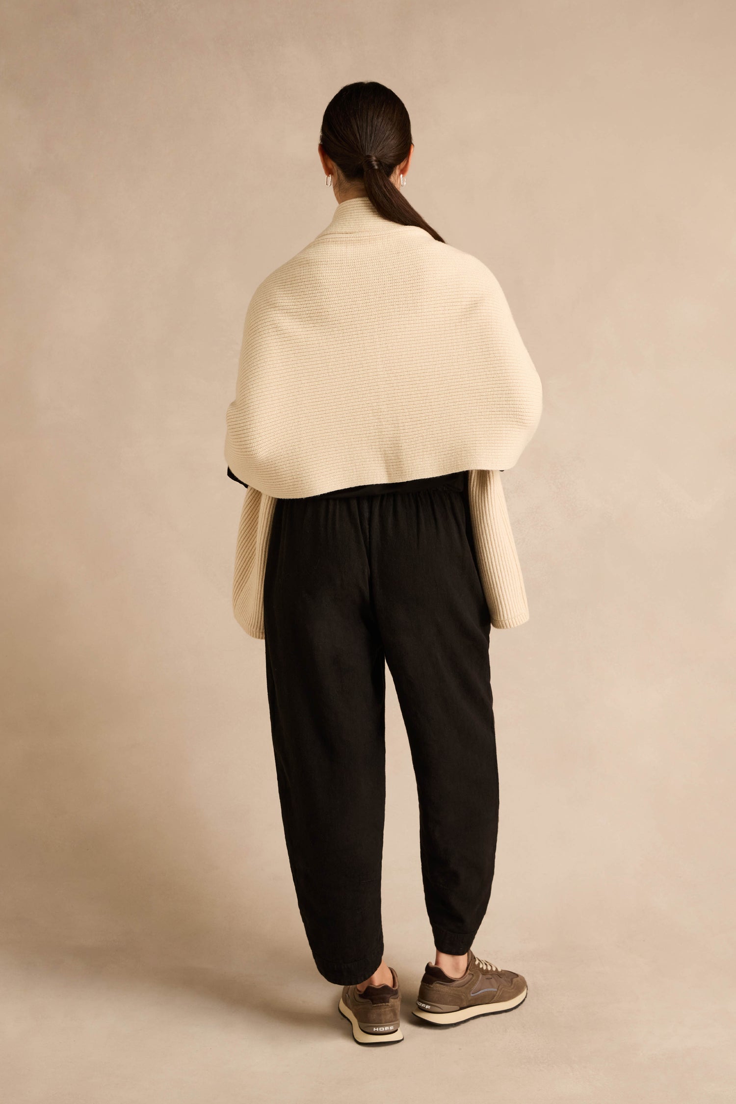 A person stands facing away against a neutral beige background, wearing the Cream Pocket Scarf—a cream textured shawl crafted from a recycled fibre blend—paired with black loose pants, brown sneakers, and their hair tied back.