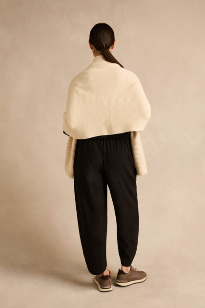 A person stands facing away against a neutral beige background, wearing the Cream Pocket Scarf—a cream textured shawl crafted from a recycled fibre blend—paired with black loose pants, brown sneakers, and their hair tied back.