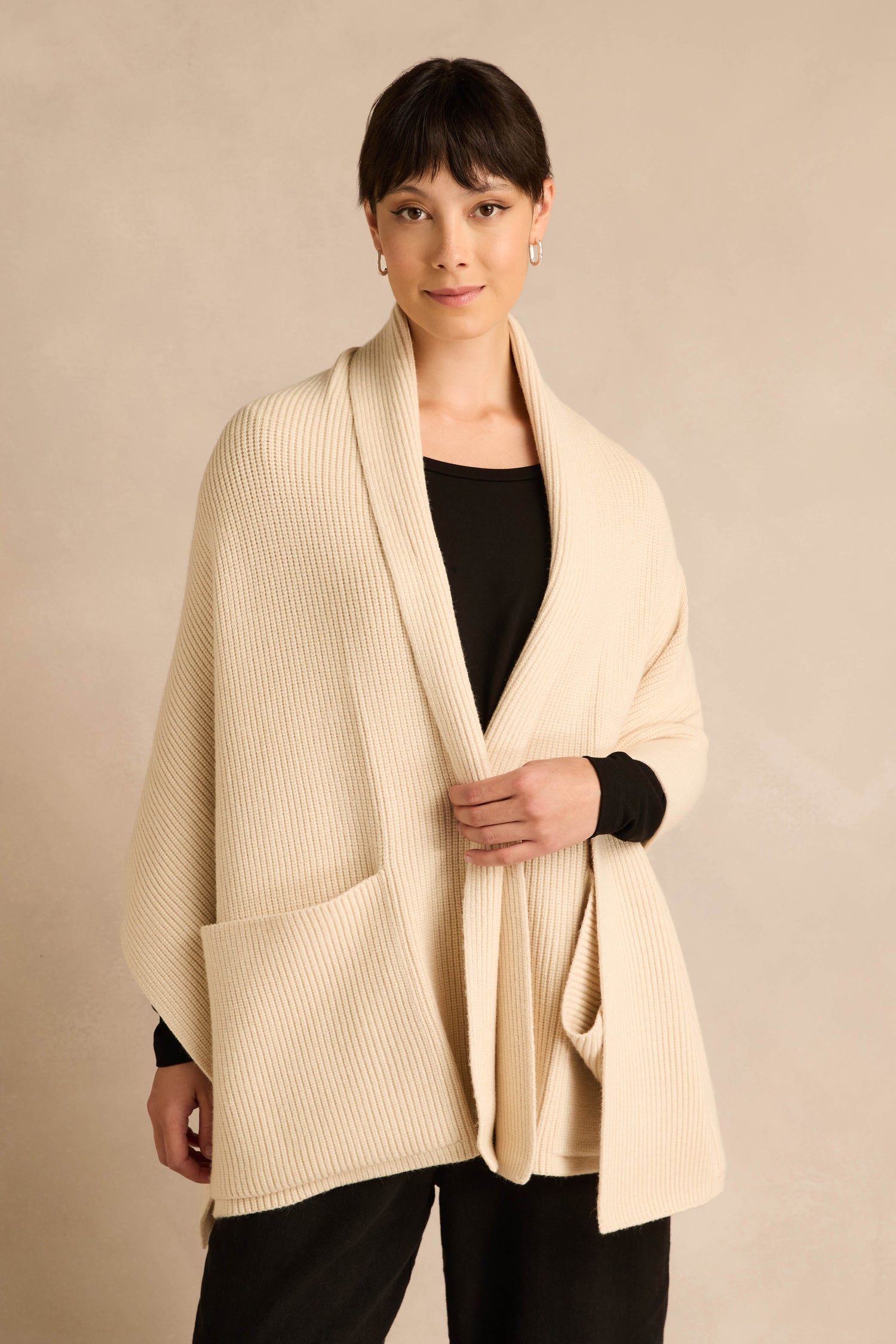 A woman stands against a neutral background wearing the Cream Pocket Scarf—a cream-colored, ribbed knit shawl made from a recycled fibre blend with two oversized pockets—layered over a black top and pants.