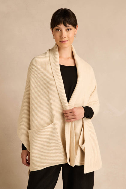 A woman stands against a neutral background wearing the Cream Pocket Scarf—a cream-colored, ribbed knit shawl made from a recycled fibre blend with two oversized pockets—layered over a black top and pants.