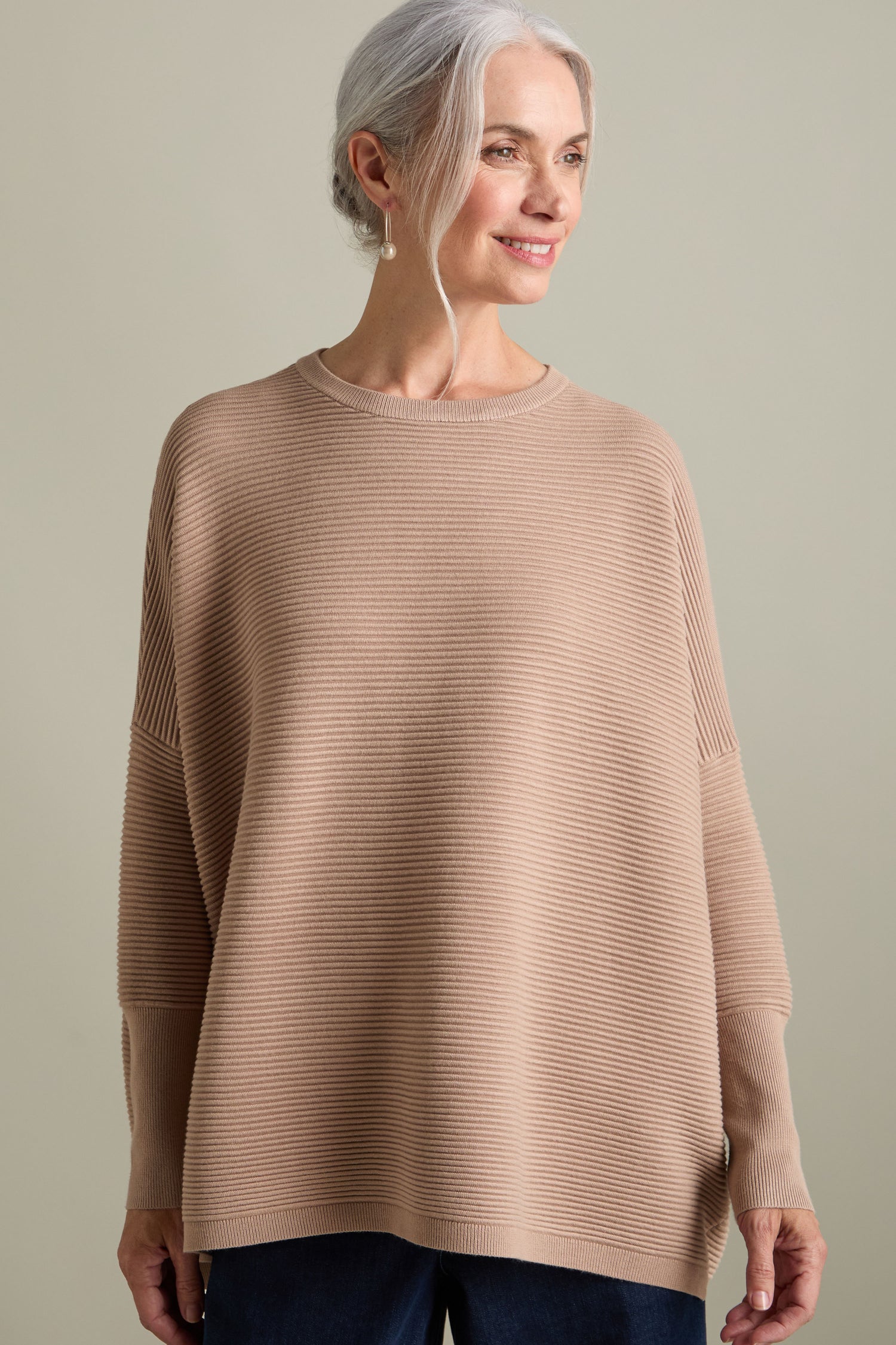 A woman with gray hair is wearing a light brown Ribbed Jumper by Paisie and dark pants, standing against a neutral background while smiling. The luxurious feel of the new-season colors adds an elegant touch to her outfit.