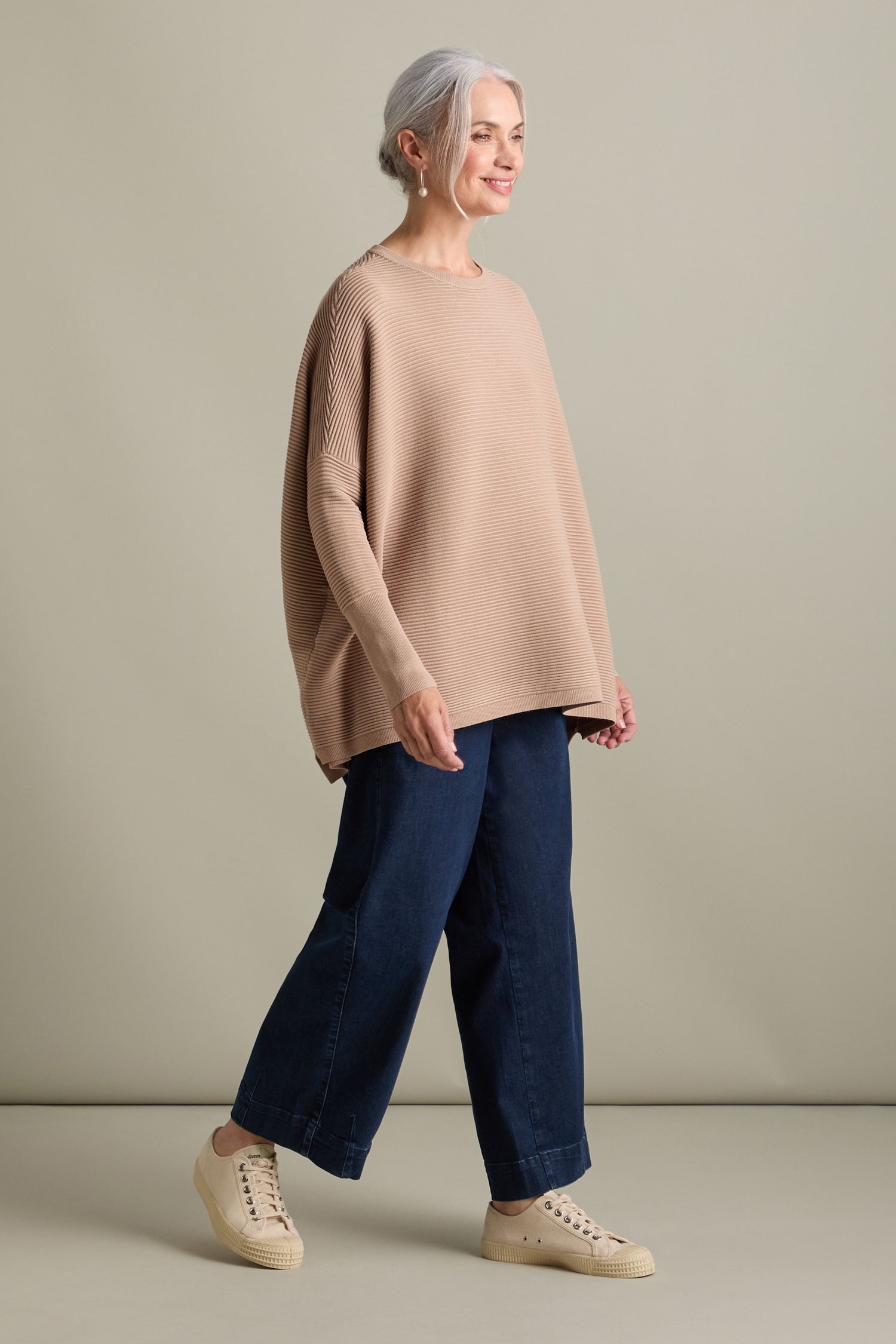 An older adult with gray hair stands against a plain background, wearing a loose beige Ribbed Jumper from Paisie, wide-leg blue jeans, and light-colored sneakers. They are looking to the side and smiling, exuding a luxurious feel in timeless new-season colors.