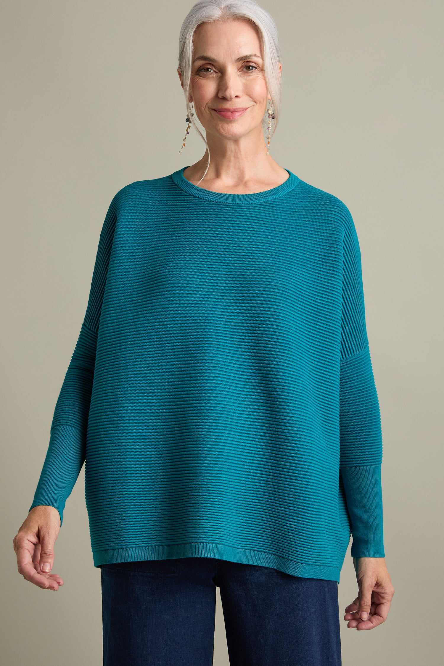 An older woman with gray hair, a luxurious feel teal Paisie Ribbed Jumper, and jeans smiles while standing against a plain background.