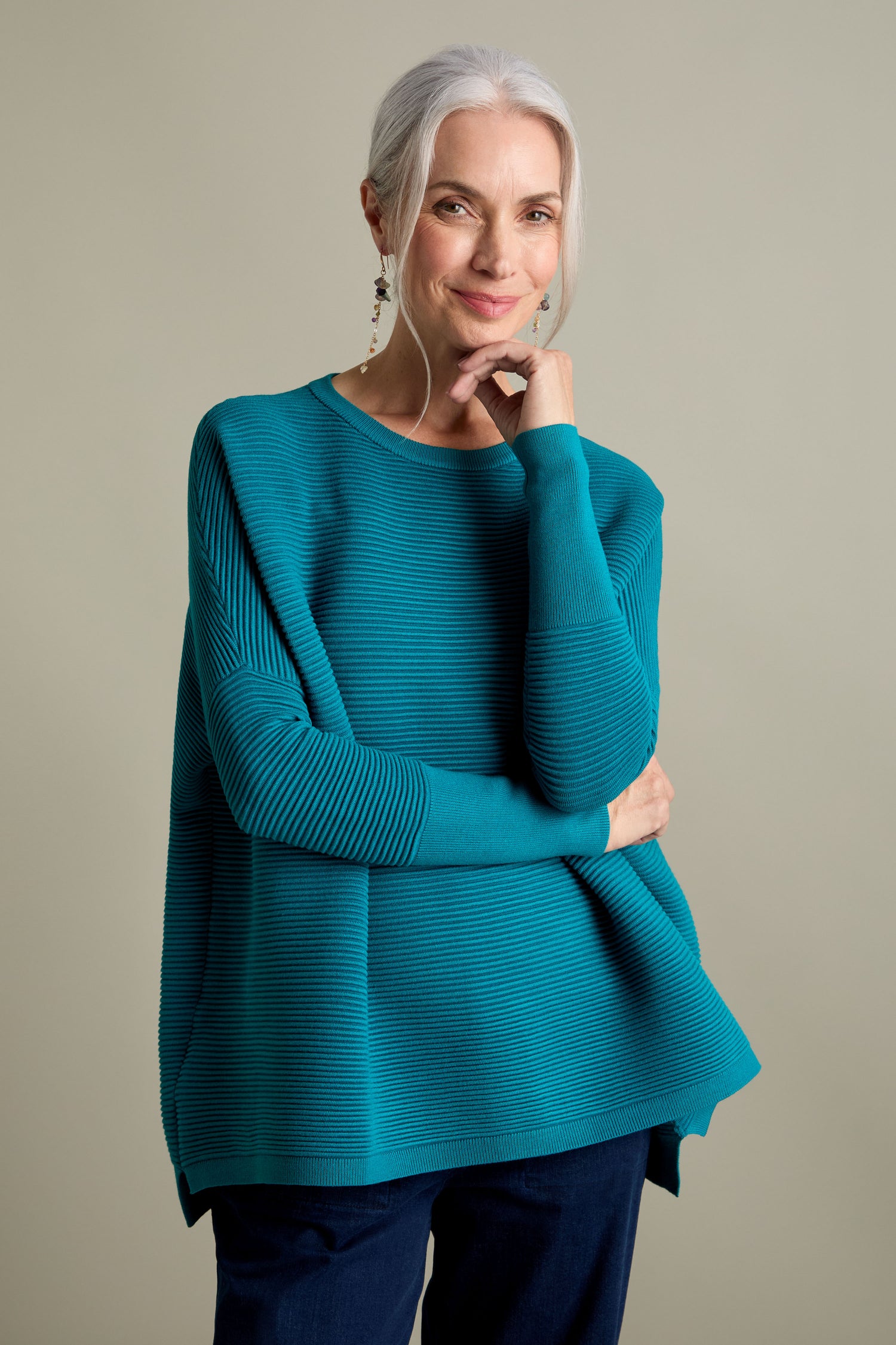 A person with long, gray hair tied back is wearing a teal Ribbed Jumper from Paisie paired with dark pants, exuding a luxurious feel while posing with one hand resting on their chin.