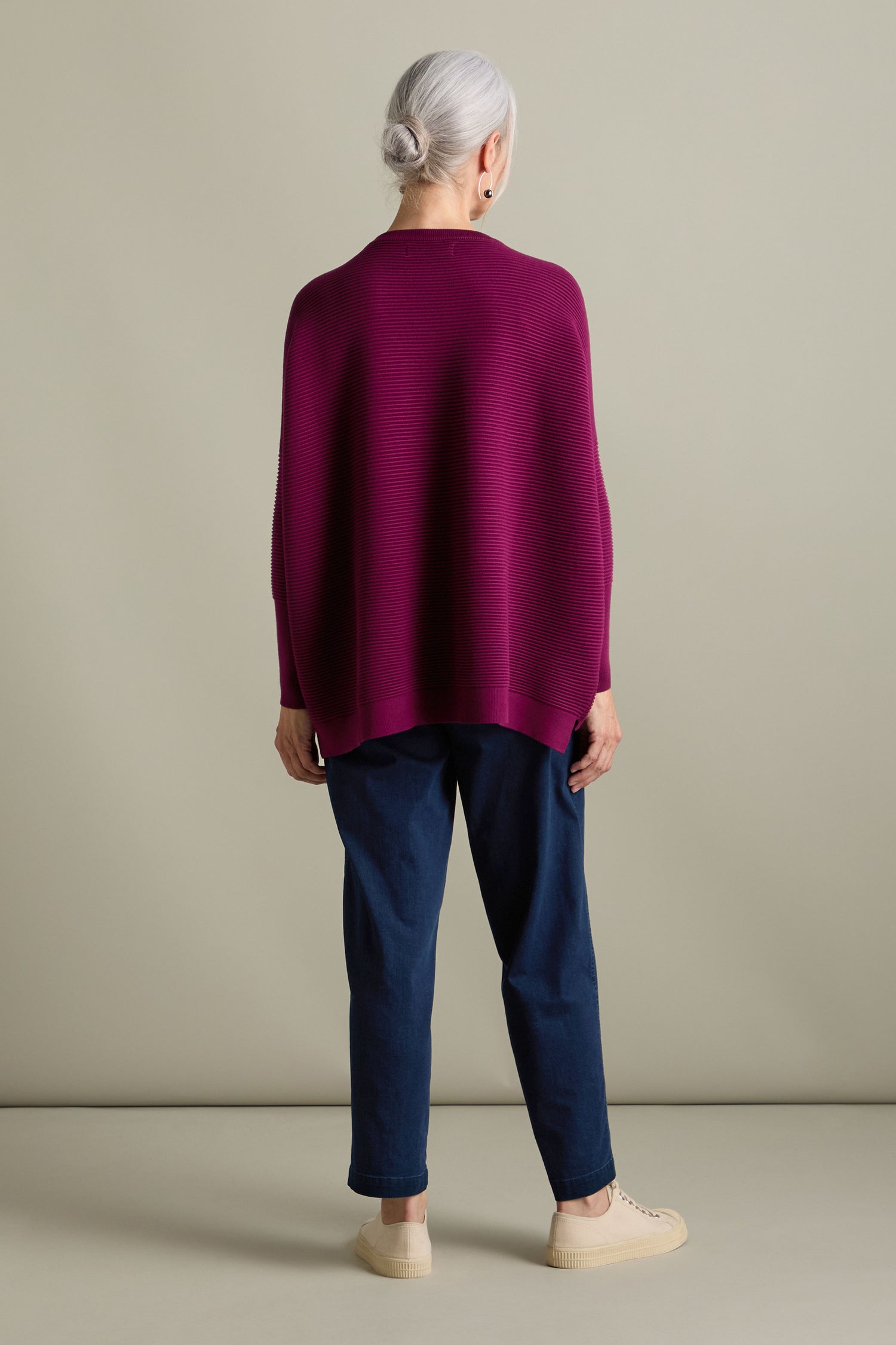 A person with grey hair in a bun, wearing a Ribbed Jumper from Paisie's new-season collection, blue pants, and beige shoes, stands facing away against a plain background.