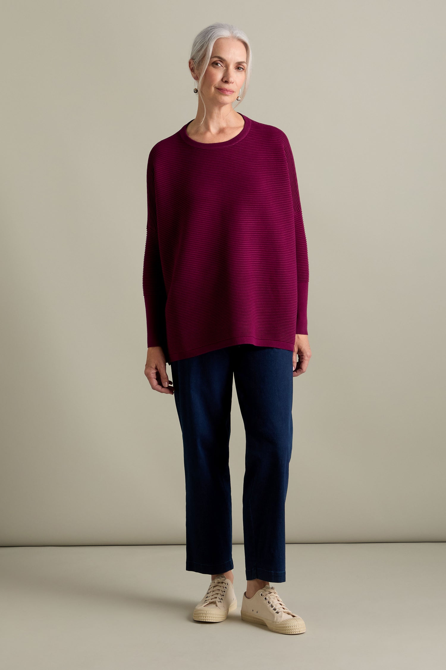 An older woman stands against a plain background, wearing a Ribbed Jumper in new-season colors, dark pants, and beige sneakers. She has short gray hair and a neutral expression.