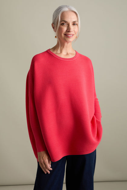 An older woman with white hair tied back is wearing a loose, bright red Ribbed Jumper from Paisie and dark pants. The jumper's new-season colors give it a luxurious feel as she stands against a plain background.