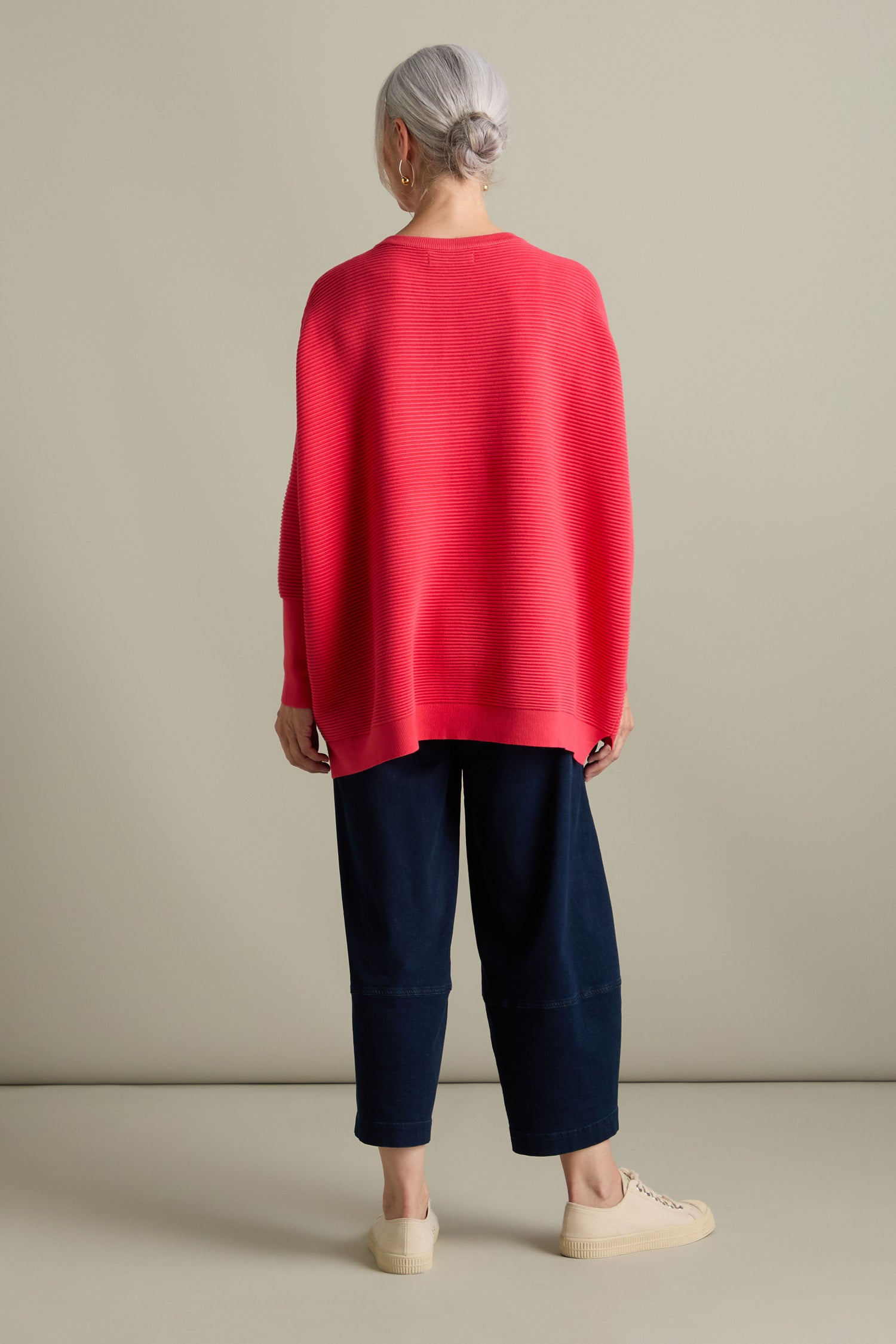 Seen from behind, a person with gray hair is wearing a loose-fitting Ribbed Jumper in red by Paisie, paired with dark blue pants and beige shoes. The luxurious feel of the jumper and its new-season colors stand out against the plain background.