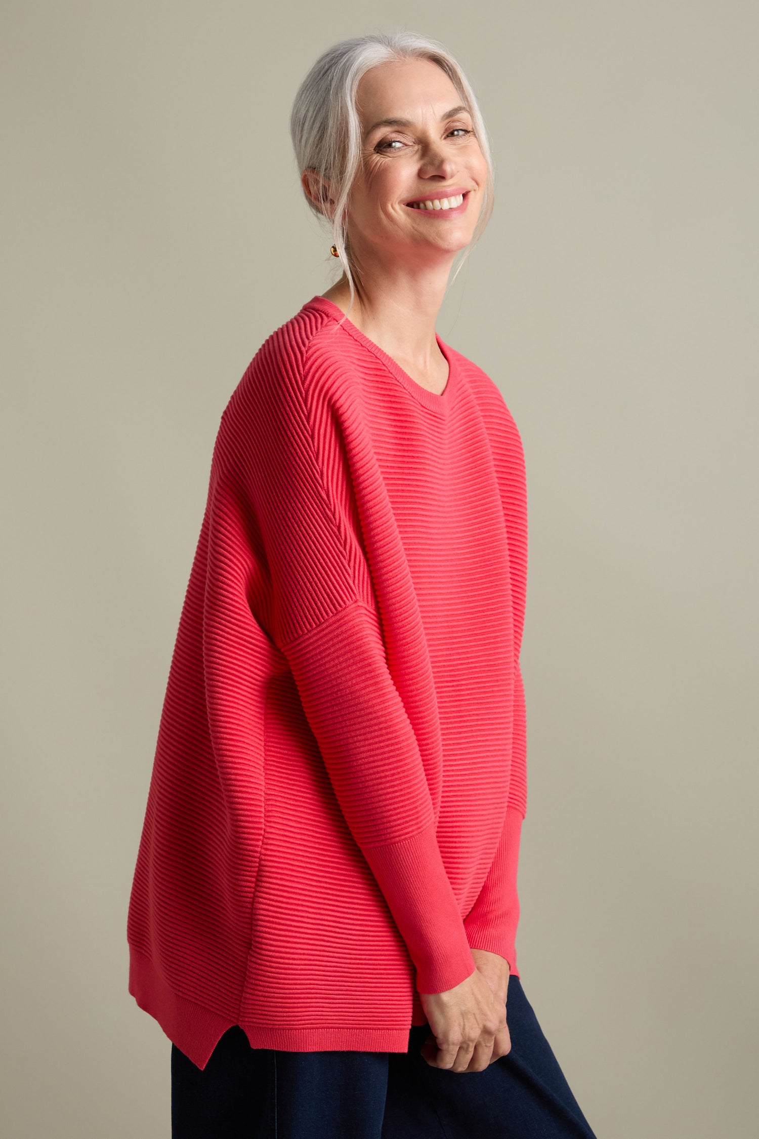 A smiling person with gray hair, draped in a loose-fitting, luxurious red Ribbed Jumper from Paisie, stands against a plain background.