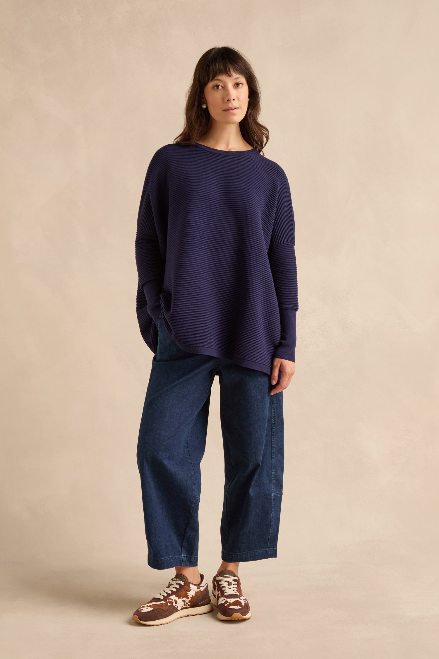 A woman stands against a beige background, wearing the Navy Ribbed Jumper, wide-leg denim jeans, and brown sneakers.