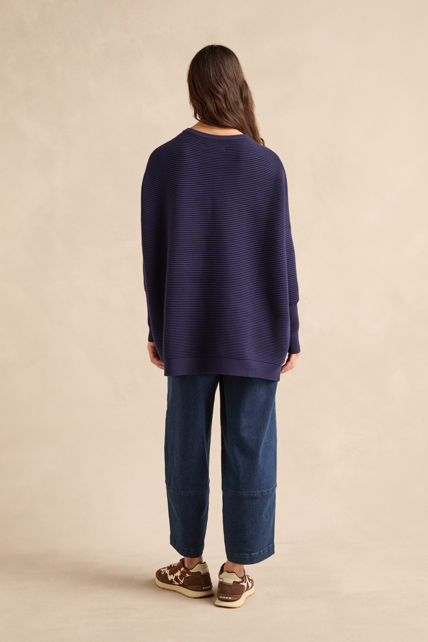 Person standing against a beige background, facing away, wearing a Navy Ribbed Jumper in a soft viscose blend, paired with wide-leg denim pants and brown sneakers.