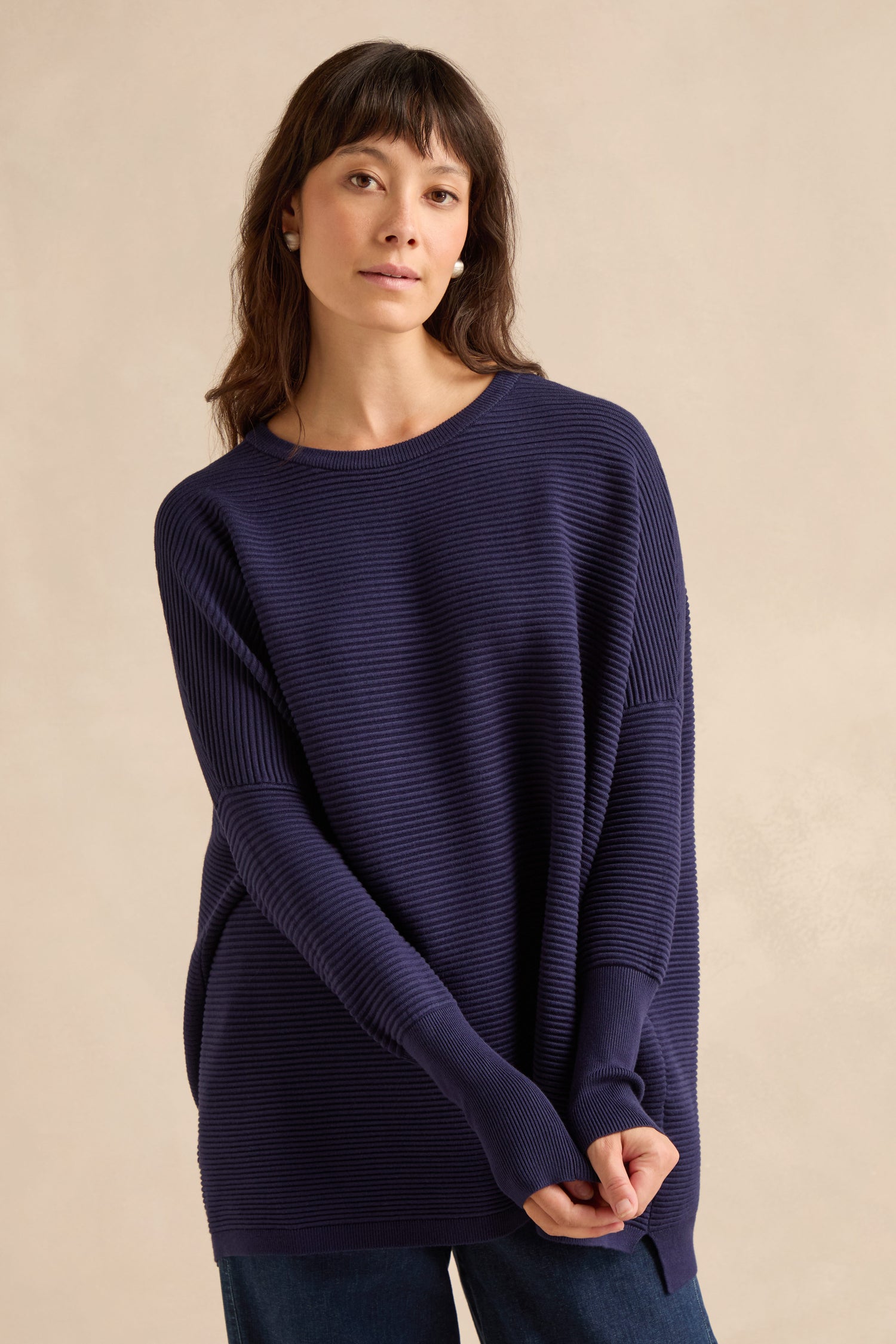 A woman with straight brown hair wears the Navy Ribbed Jumper, a loose-fitting piece crafted from a soft viscose blend, paired with blue jeans as she stands against a plain beige background.