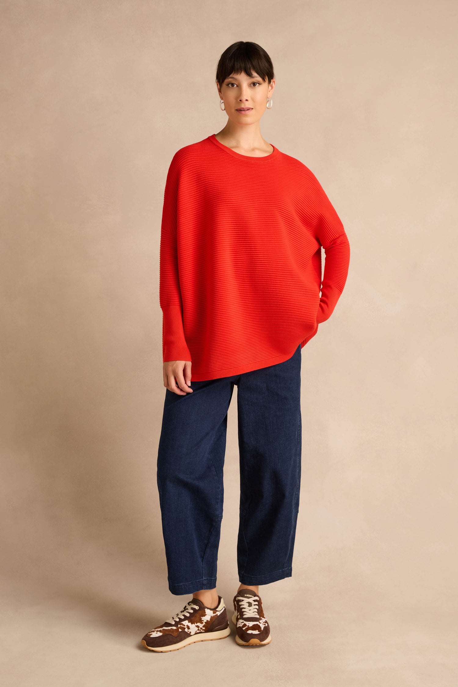 A woman stands against a beige background wearing a Red Ribbed Jumper, wide-leg dark blue jeans, and brown sneakers.
