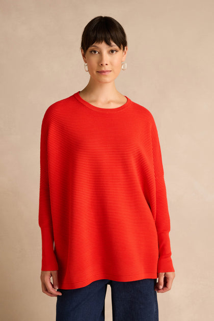 A woman with short dark hair and silver hoop earrings stands against a neutral background, wearing the Red Ribbed Jumper—a bright red, long-sleeve piece crafted from a soft viscose blend—paired with dark pants.