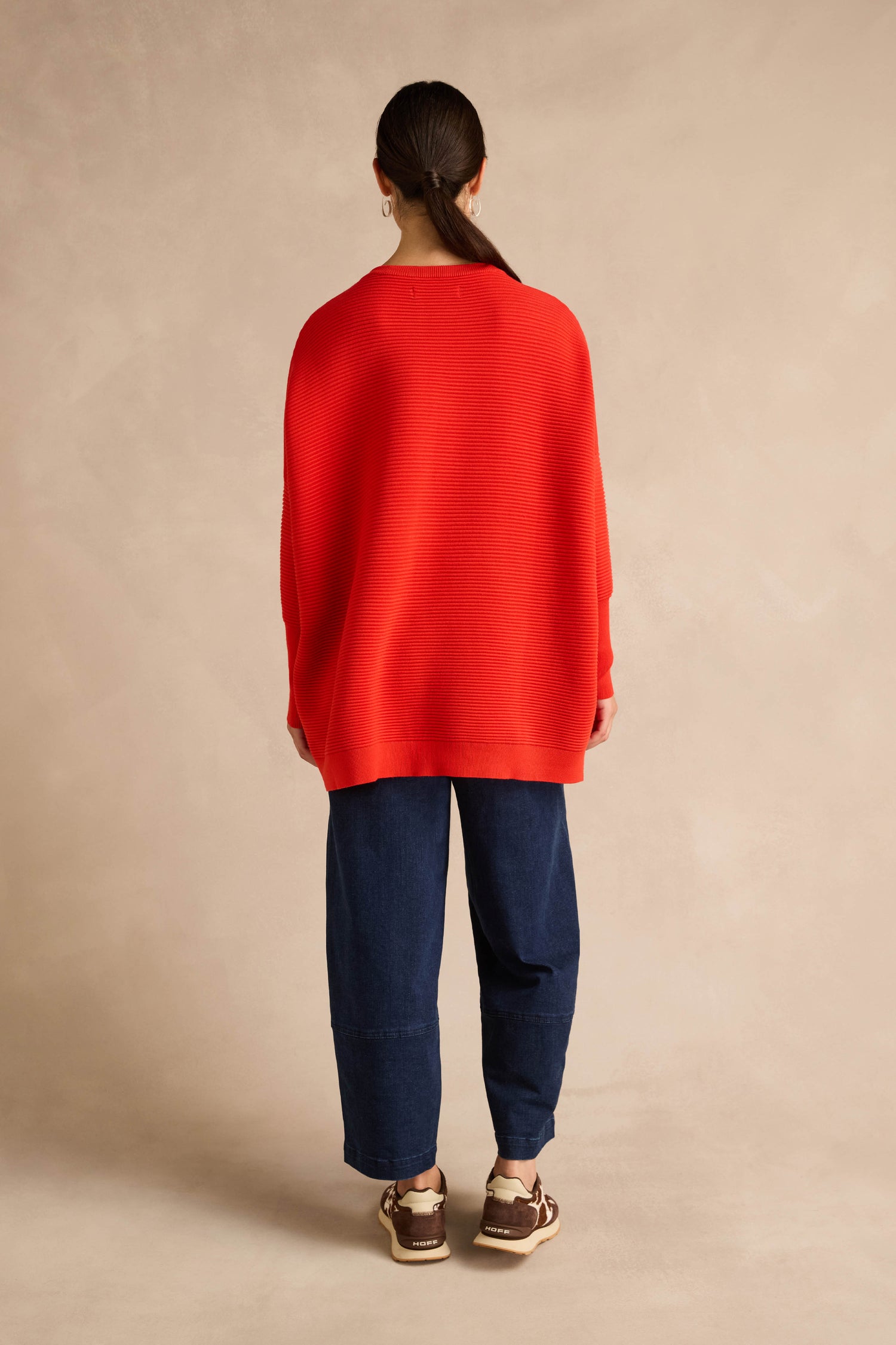 A person stands facing away against a plain beige background, wearing a loose Red Ribbed Jumper, dark blue wide-leg pants, and brown sandals.