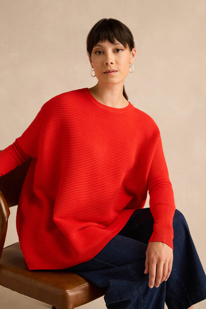A woman wears the Red Ribbed Jumper styled loosely with blue jeans as she sits on a brown chair against a neutral background, gazing at the camera with a neutral expression.
