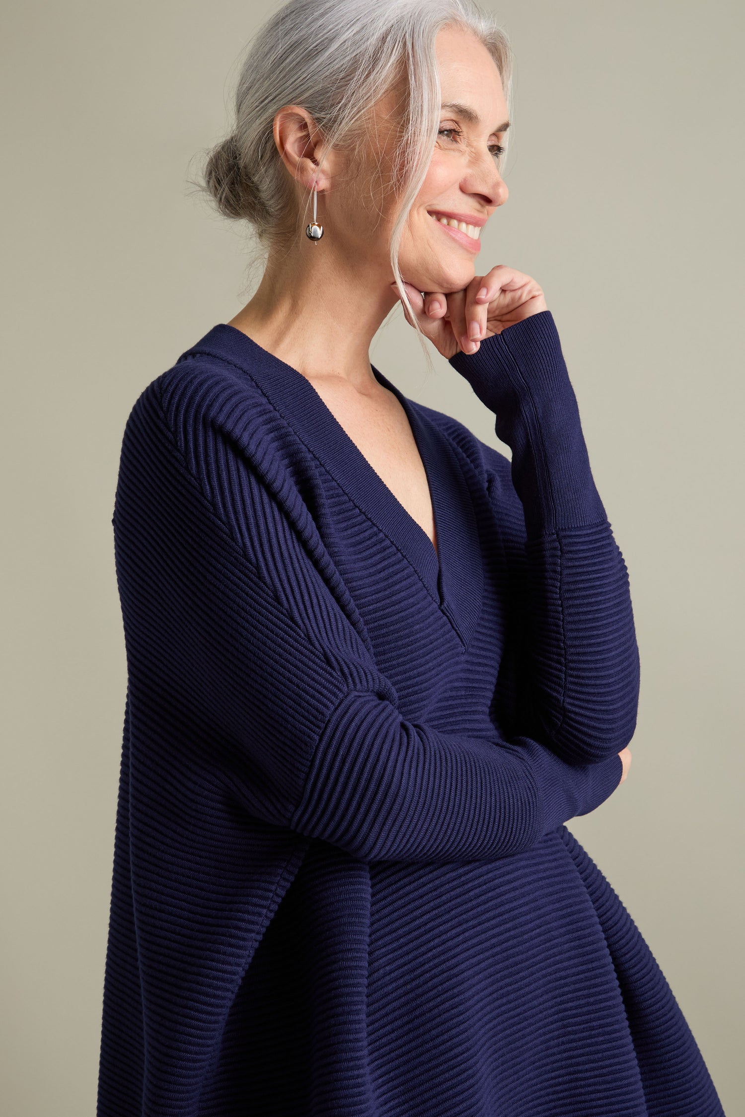 A woman with gray hair pulled back is smiling, wearing a luxurious V Neck Ribbed Jumper in navy blue. She is posing sideways with her chin resting on her hand.