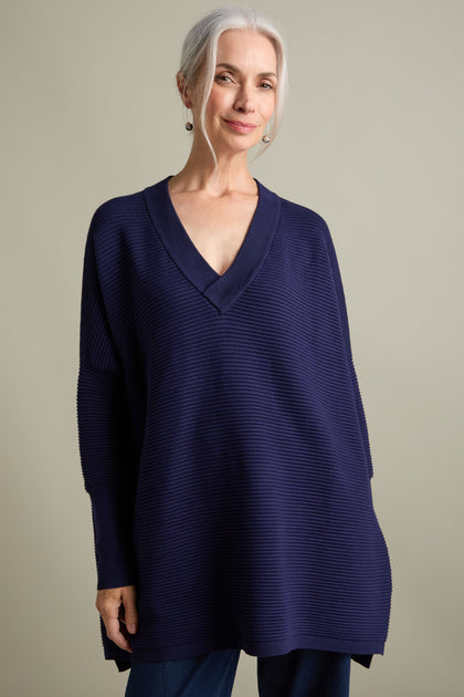 An older adult with silver hair tied back, wearing a sophisticated V Neck Ribbed Jumper from Paisie that showcases a luxurious ribbed knit design, stands against a plain background.