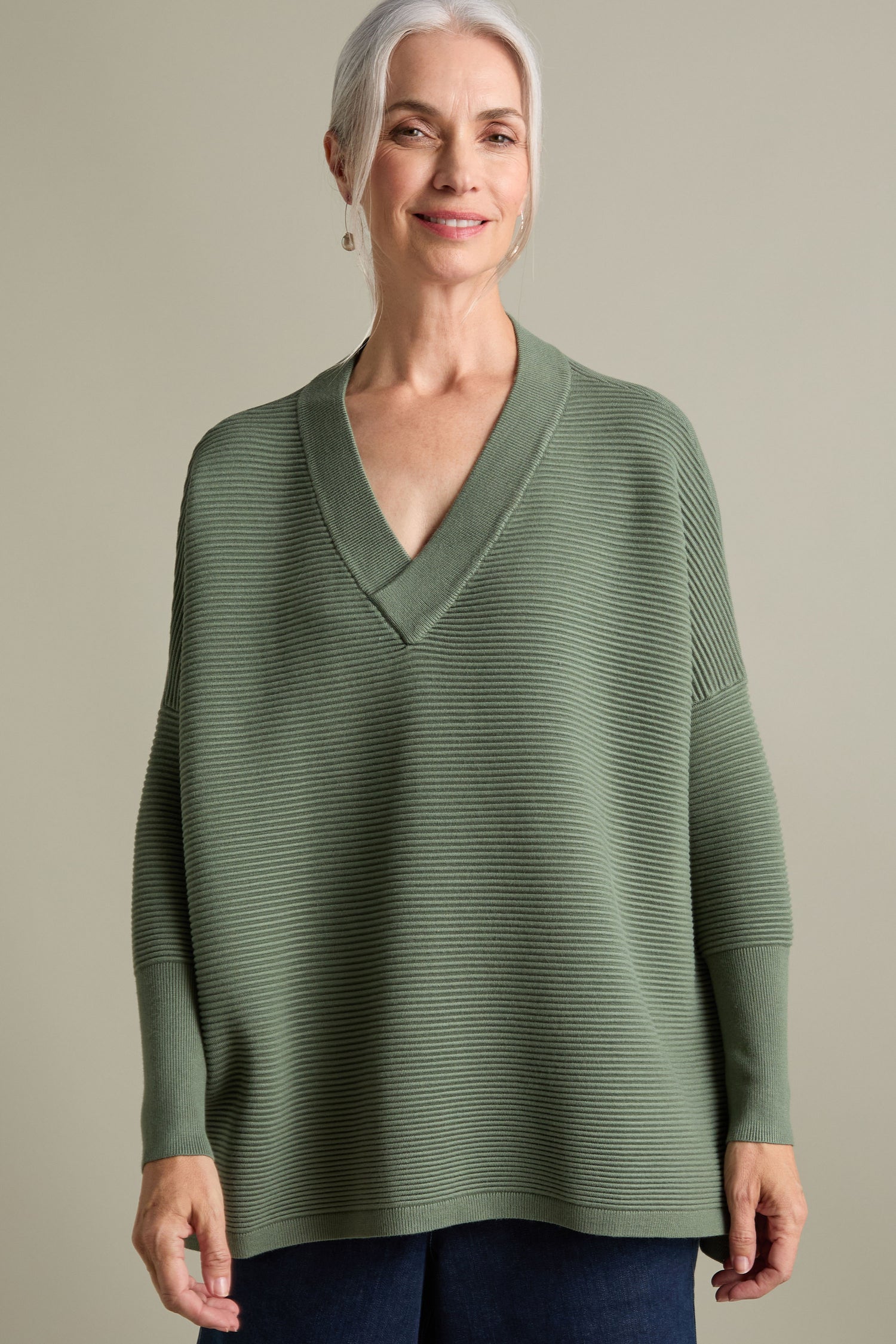 A smiling woman with gray hair wears a luxurious olive green V Neck Ribbed Jumper and dark pants against a plain background.