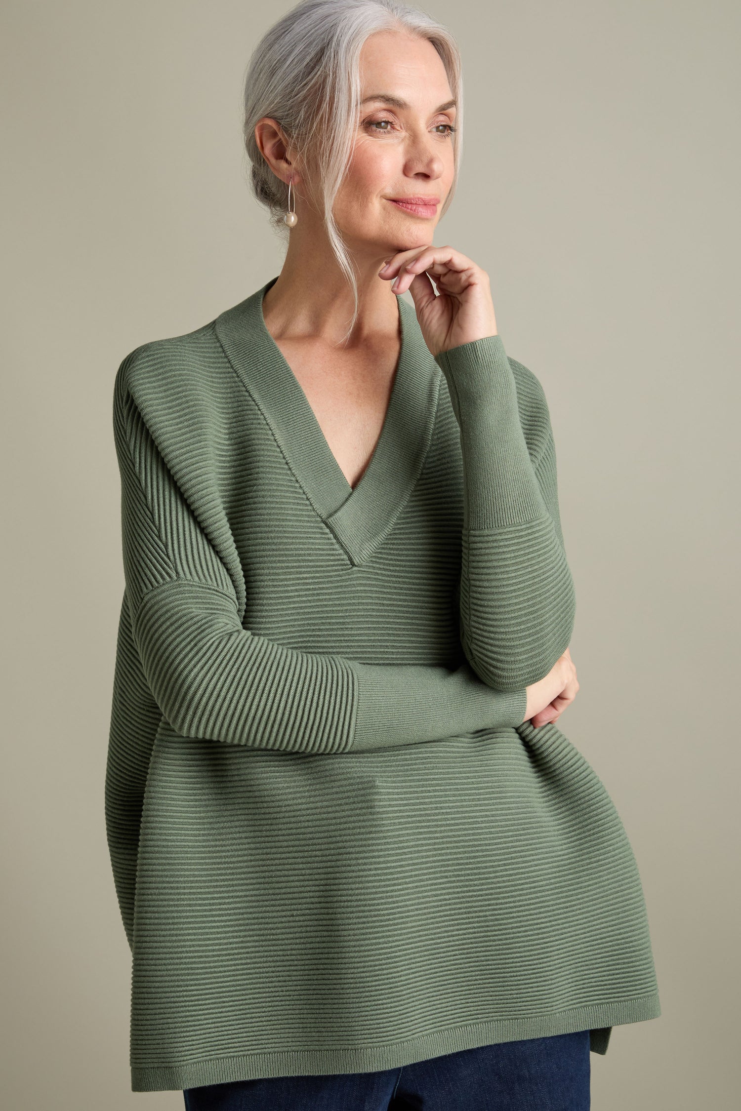 A woman with gray hair, wearing a luxurious V Neck Ribbed Jumper in green, stands with her arms crossed and one hand touching her chin against a beige background.