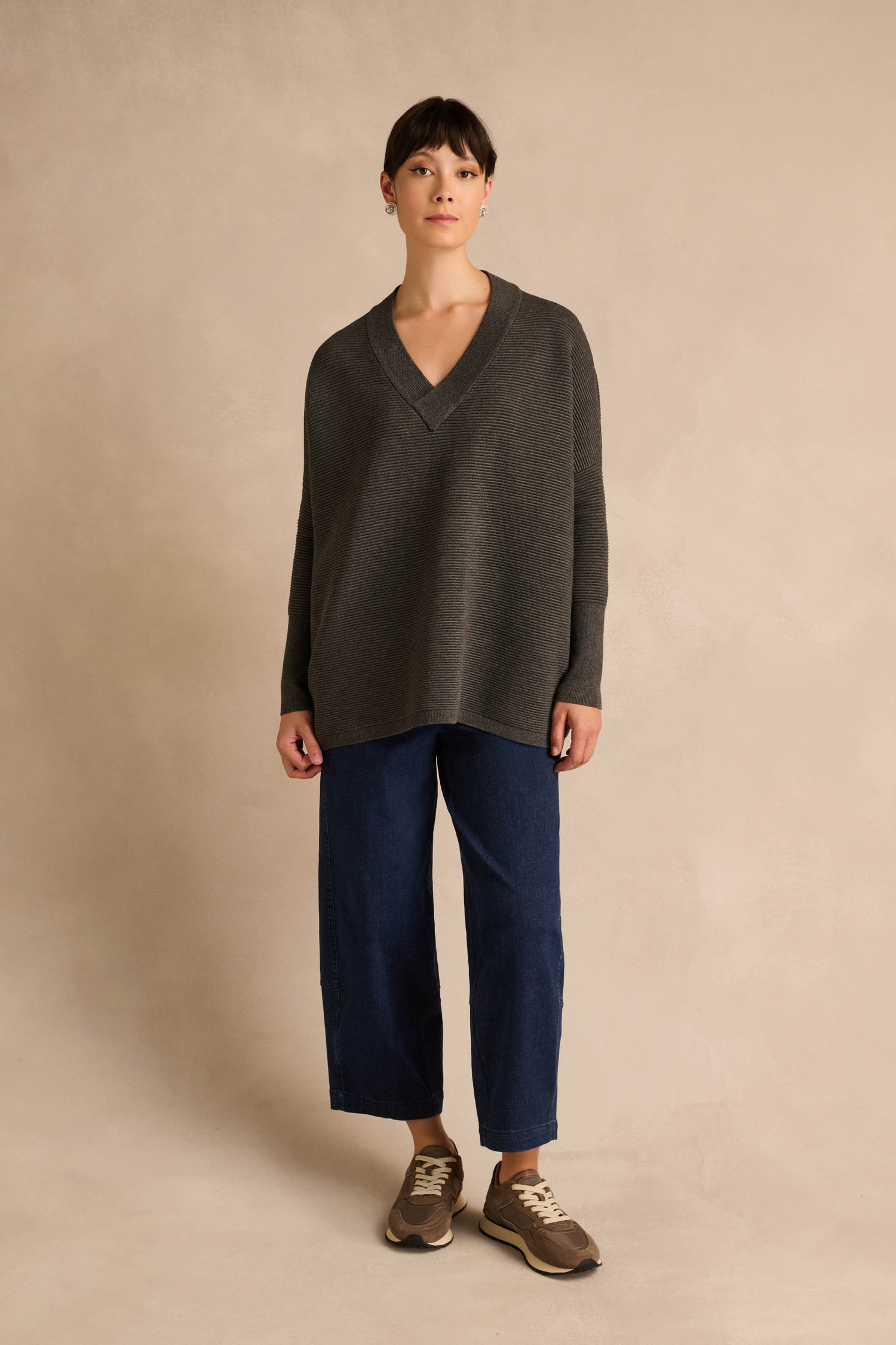 A person stands against a plain beige background wearing a relaxed fit Charcoal V Neck Ribbed Jumper, crafted from a soft viscose blend, paired with wide-leg blue jeans and brown sneakers.
