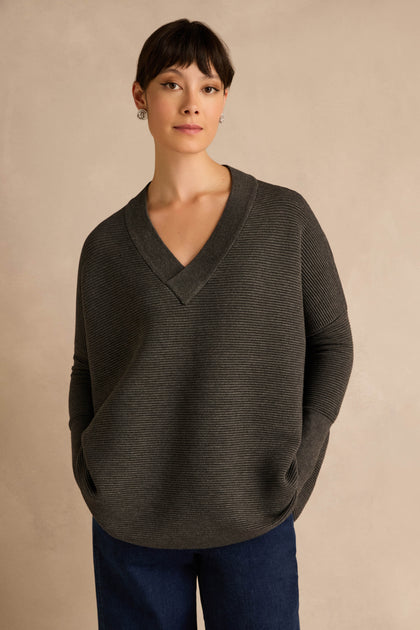 A person wearing the Charcoal V Neck Ribbed Jumper, made from a soft viscose blend, stands against a plain beige background with their hands in their pockets.