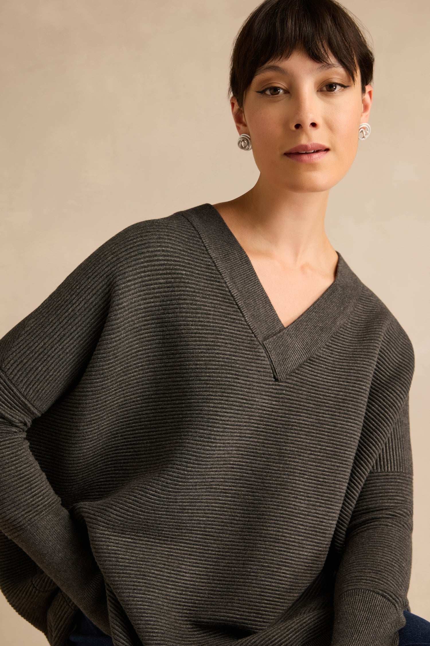 A woman wears the Charcoal V Neck Ribbed Jumper, designed with long sleeves and crafted from a soft viscose blend for enhanced comfort. Featuring a relaxed fit, she stands against a neutral background, looking directly at the camera.