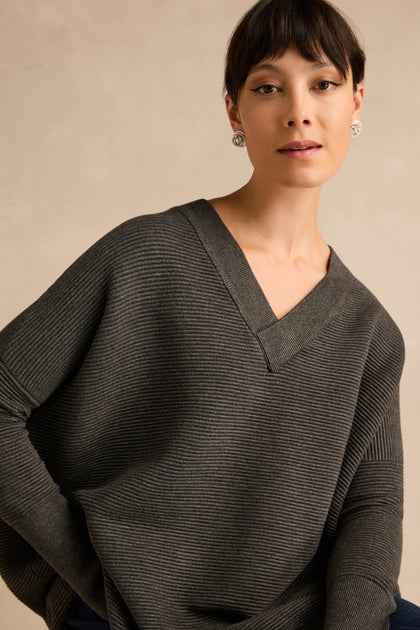 A woman wears the Charcoal V Neck Ribbed Jumper, designed with long sleeves and crafted from a soft viscose blend for enhanced comfort. Featuring a relaxed fit, she stands against a neutral background, looking directly at the camera.