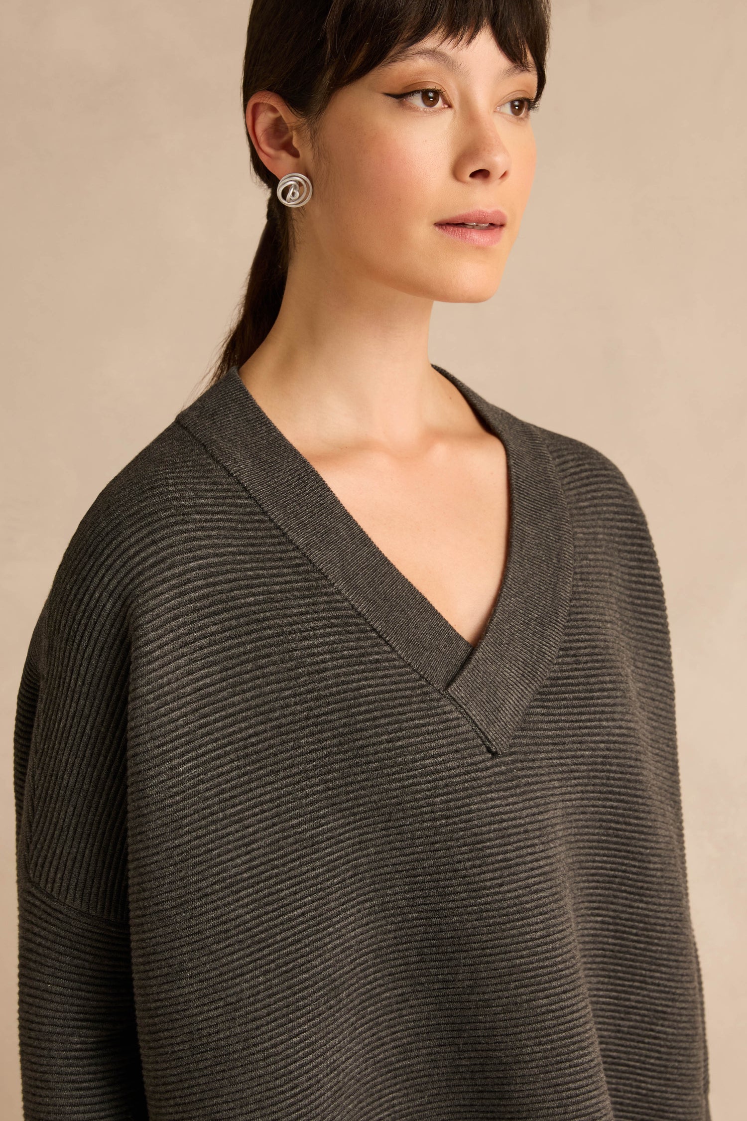 A woman wearing the Charcoal V Neck Ribbed Jumper, crafted from a soft viscose blend, paired with silver swirl earrings, stands against a neutral background.