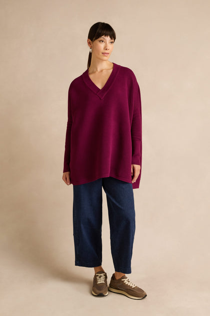 A woman stands against a neutral background wearing a relaxed fit Wine V Neck Ribbed Jumper, paired with wide-leg blue jeans and brown sneakers. The soft viscose blend of the sweater adds comfort and ease to her laid-back look.