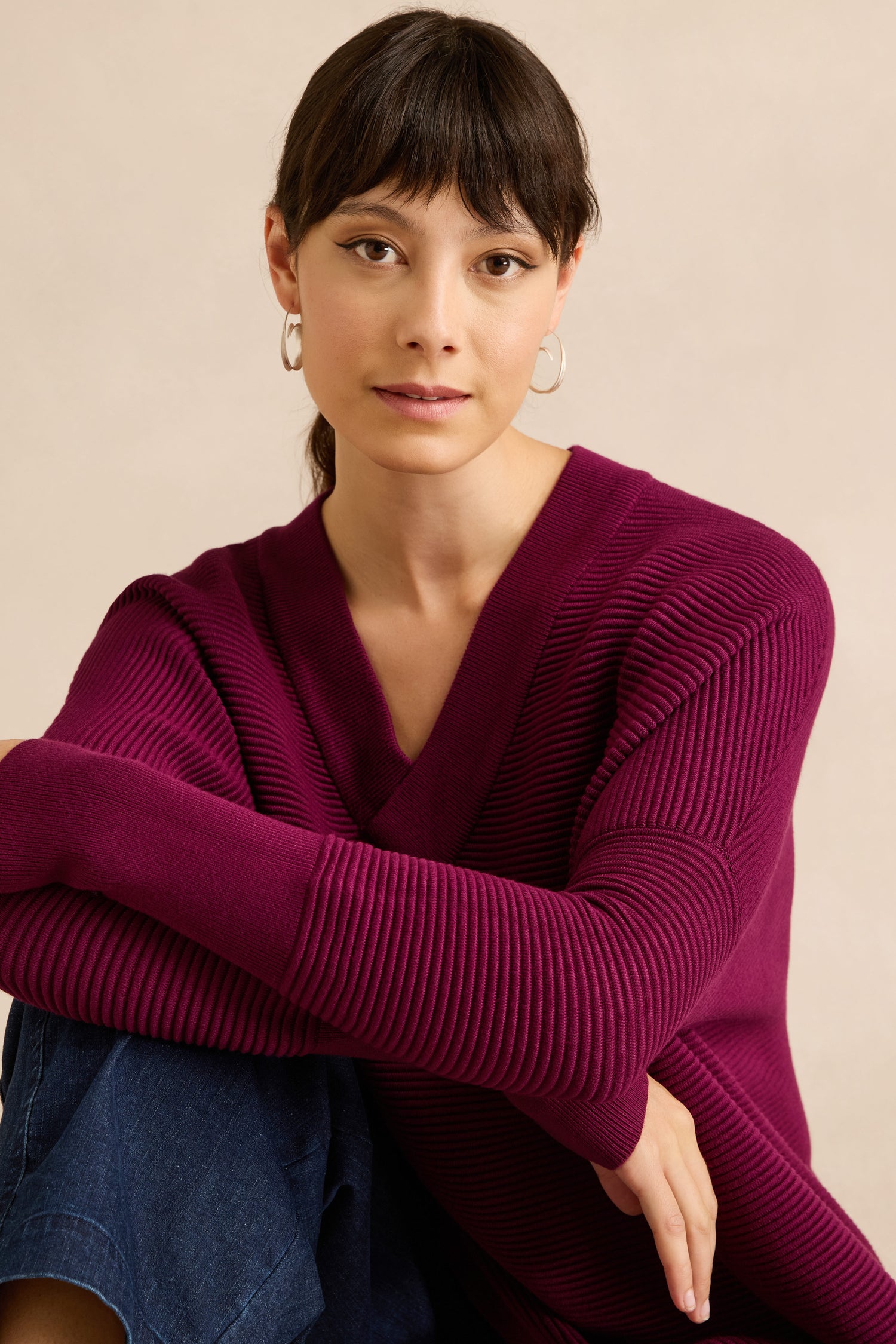 A woman with brown hair and hoop earrings wears the Wine V Neck Ribbed Jumper as she sits with her arms crossed over her knees against a neutral background. Crafted from a soft viscose blend, this relaxed fit jumper offers both comfort and style.