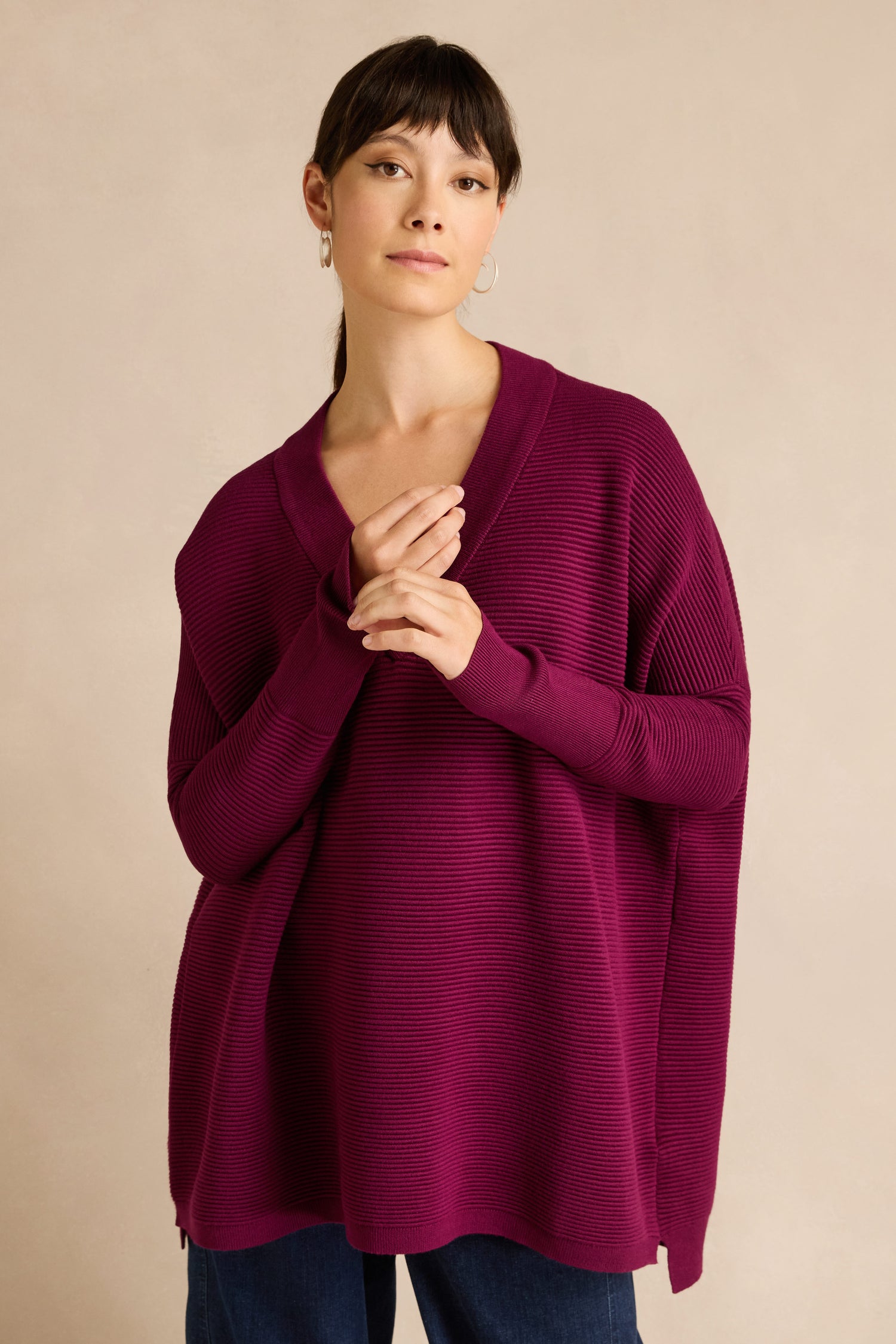 A person stands with hands clasped, wearing a relaxed fit jumper crafted from a soft viscose blend—specifically, the Wine V Neck Ribbed Jumper—paired with dark pants against a plain beige background.
