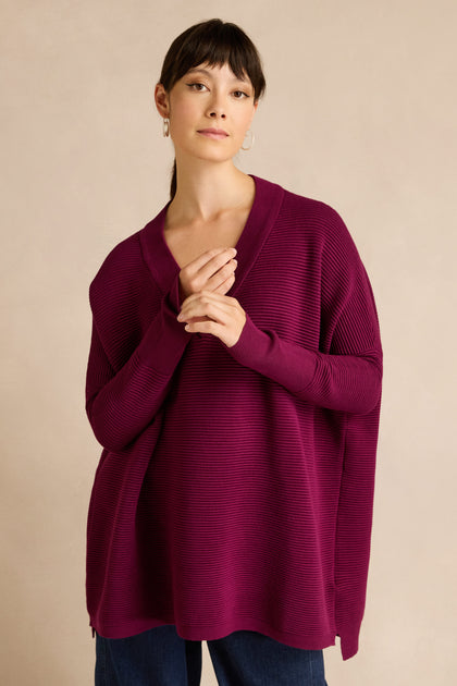 A person stands with hands clasped, wearing a relaxed fit jumper crafted from a soft viscose blend—specifically, the Wine V Neck Ribbed Jumper—paired with dark pants against a plain beige background.