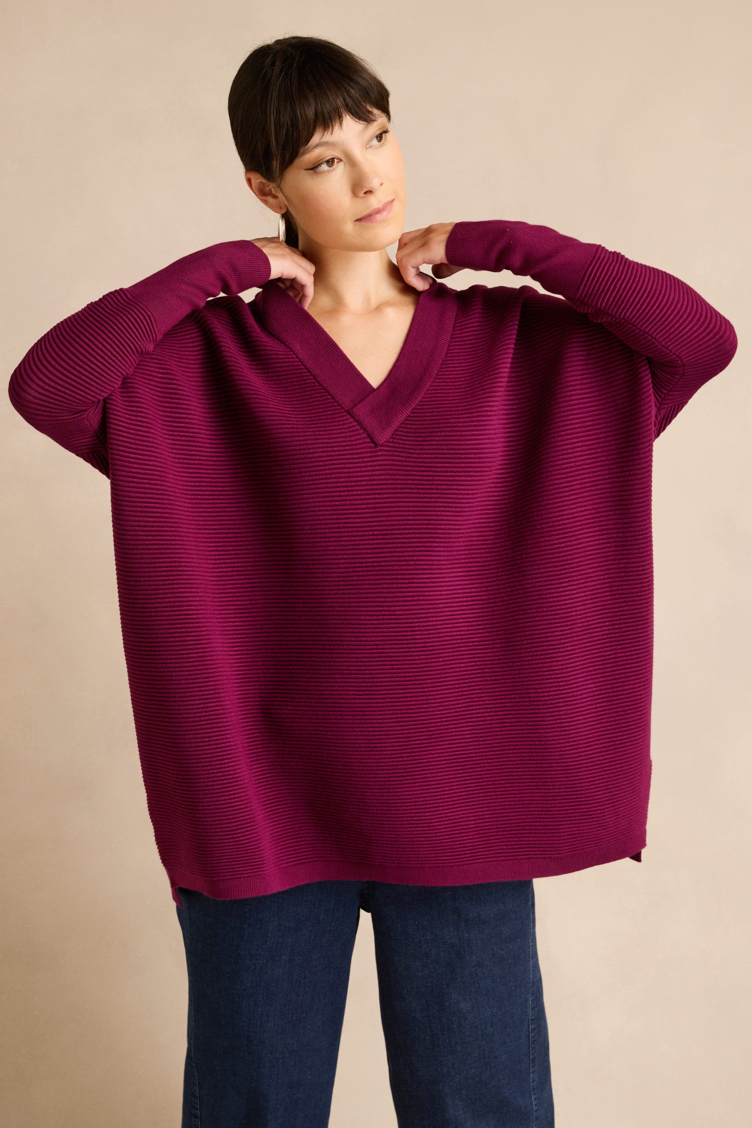 A person wearing the Wine V Neck Ribbed Jumper—a relaxed fit, soft viscose blend V-neck ribbed jumper—in magenta paired with blue jeans, standing against a plain beige background.