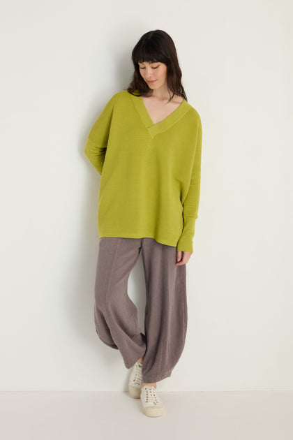 A woman stands against a white wall wearing Paisie’s V-Neck Ribbed Ochre Top, wide-legged gray pants, and white sneakers, looking down with her hands by her sides.