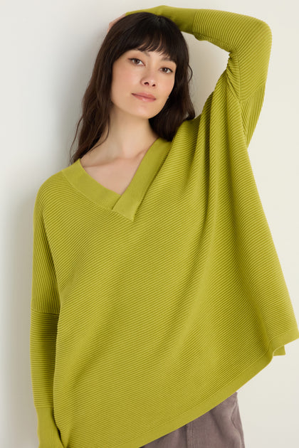 A woman with long dark hair wears a Paisie V-Neck Ribbed Ochre Top, resting one arm on her head as she stands against a plain light background.
