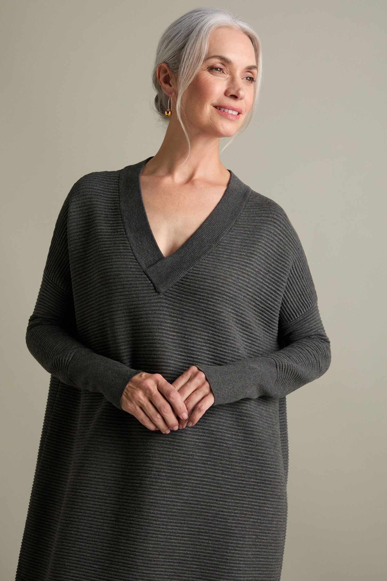 A woman with grey hair is wearing a long, dark grey V Neck Ribbed Dress that features a flattering V-neckline. She is looking slightly to her left with a smile, and her hands are clasped in front of her. The background is plain and light, giving her a timeless versatile look.