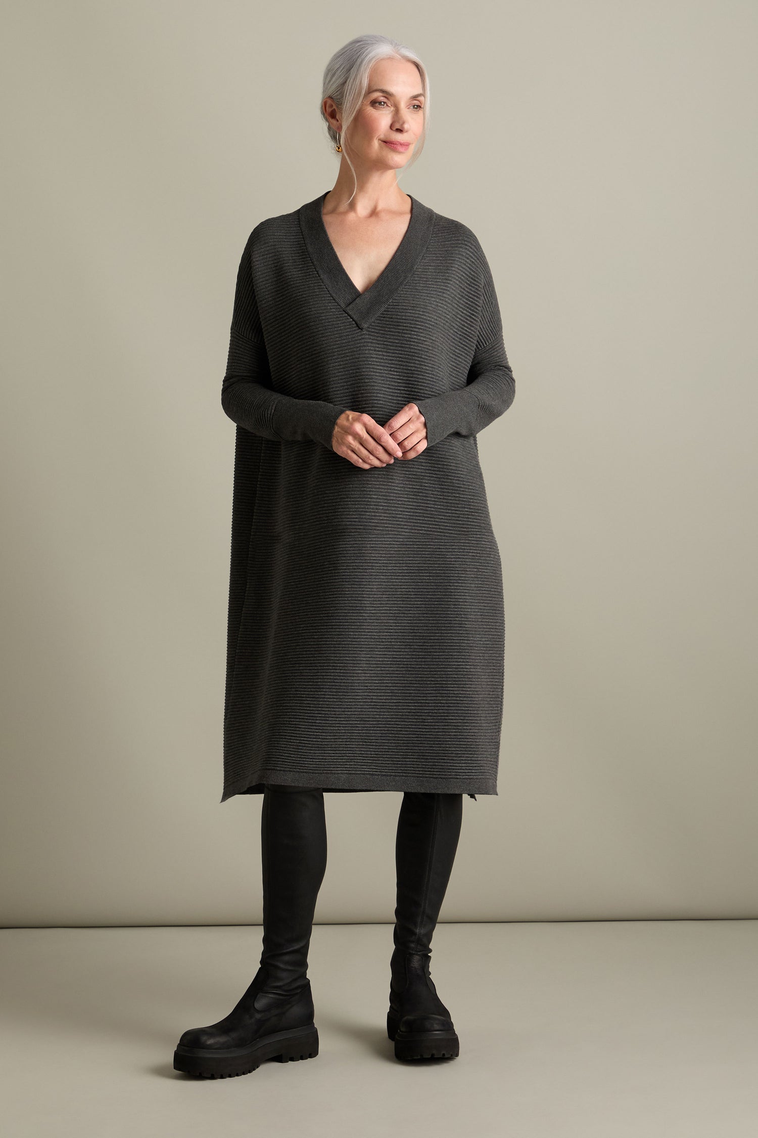 A woman with gray hair stands against a plain background wearing the V Neck Ribbed Dress, a long, dark gray knit attire featuring a flattering V-neckline, paired with black boots. She has a neutral expression, her hands clasped in front of her, embodying a timeless versatile look.