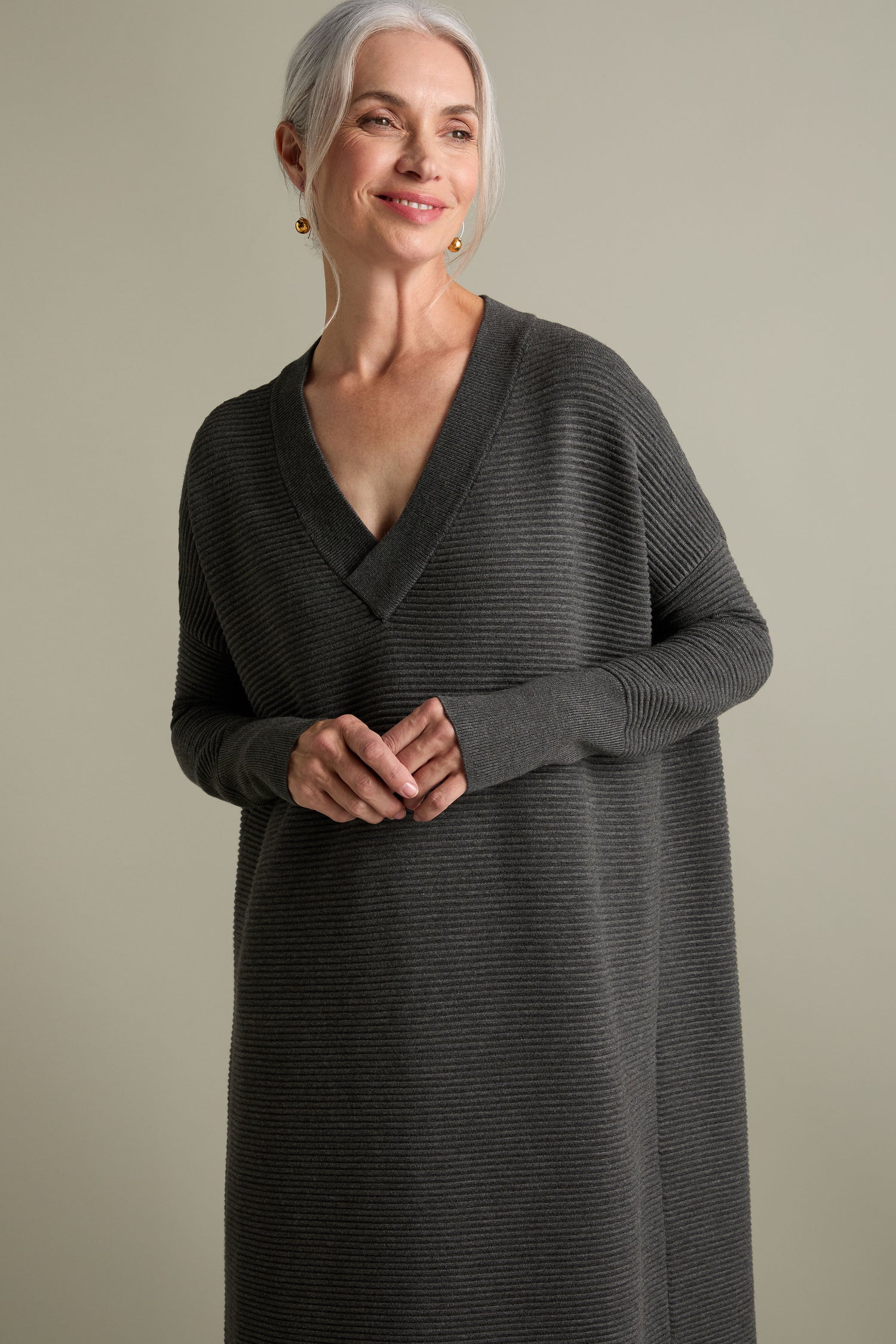 A woman with gray hair smiles while wearing a long, dark gray V Neck Ribbed Dress by Paisie and gold earrings, showcasing a timeless versatile look against a neutral background.