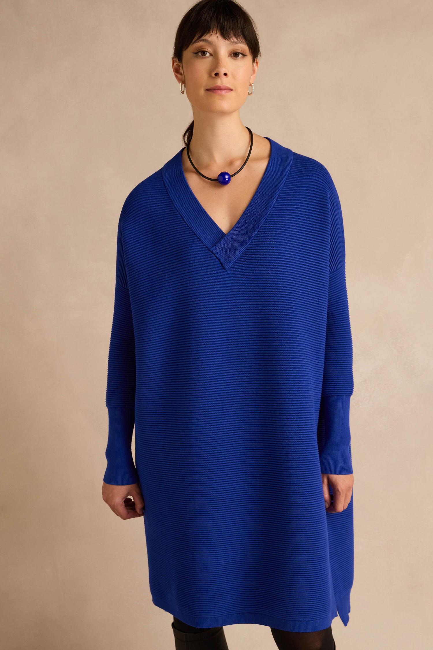 A woman stands against a beige background, wearing the V Neck Ribbed Dress in cobalt blue—a loose-fitting, long-sleeve style—paired with a black necklace featuring a blue pendant.