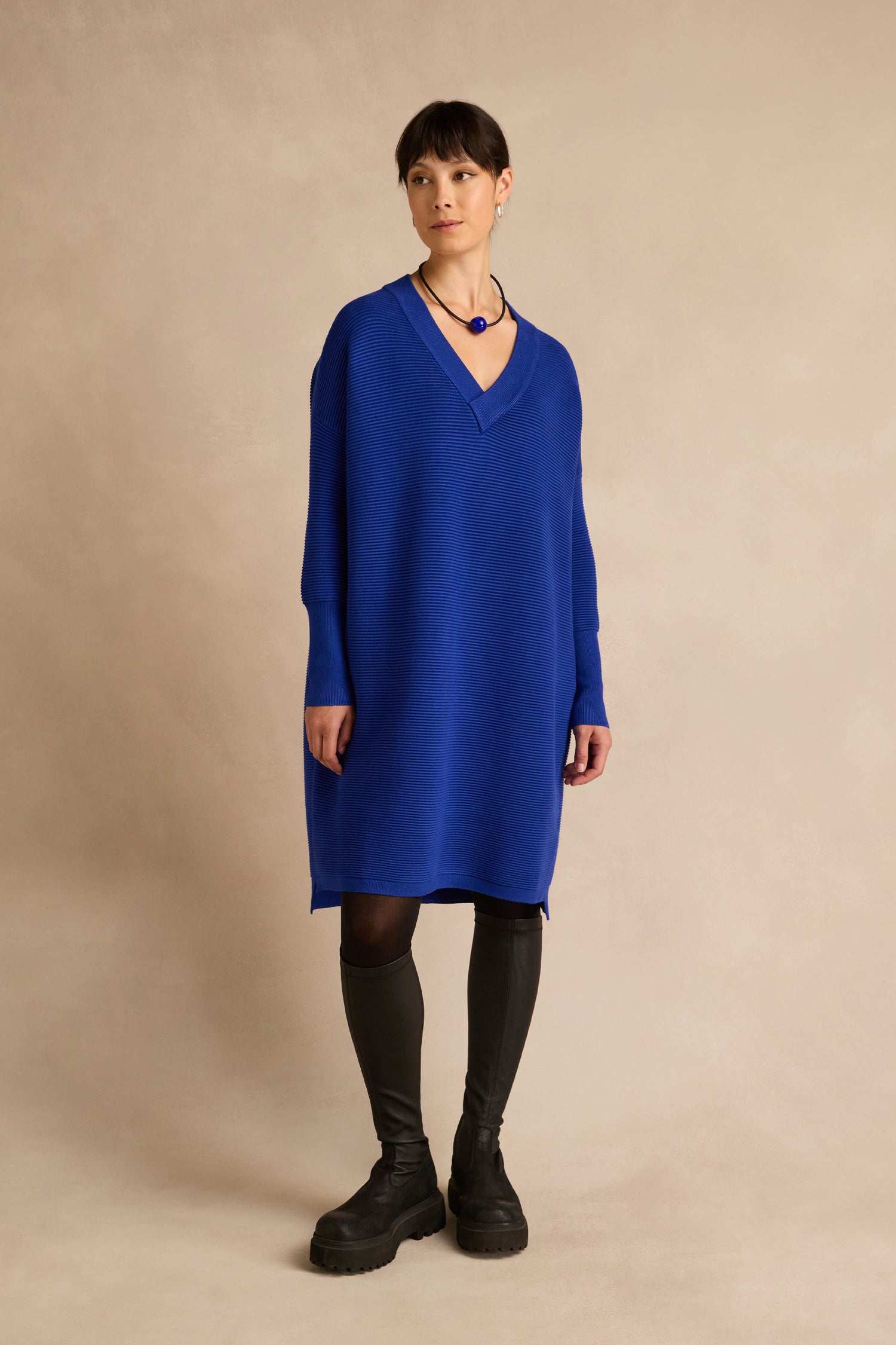 A woman stands against a neutral beige background wearing the V Neck Ribbed Dress in blue, styled with black tights and chunky black boots.