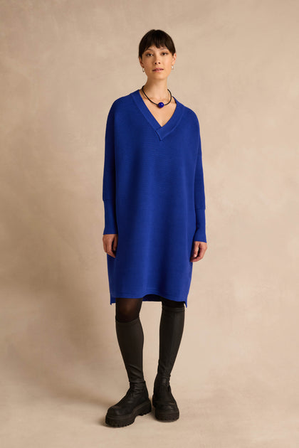 A woman stands against a beige background wearing the V Neck Ribbed Dress in blue, paired with black tights, knee-high black boots, and a blue necklace.