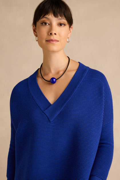 A woman is wearing the V Neck Ribbed Dress in a textured deep blue, paired with a round blue pendant necklace and small hoop earrings, as she stands against a neutral beige background.