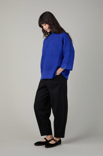 Woman standing sideways wears a loose blue Wide Sleeve Ribbed Jumper, black pants, and black Mary Jane shoes, looking down against a plain light gray background.