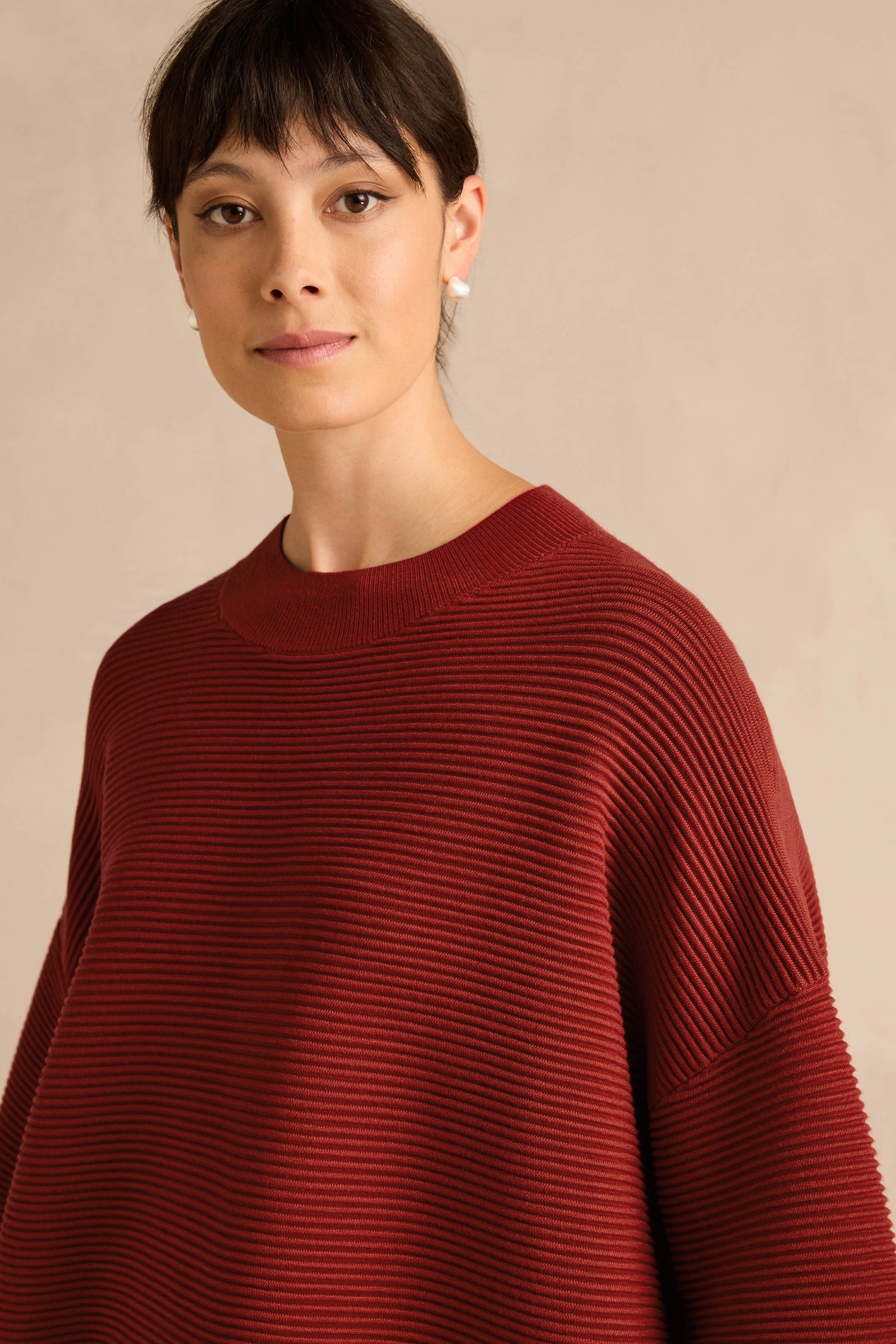 Woman with short dark hair wearing the Wide Sleeve Burgundy Ribbed Knit sweater and small earrings, standing against a plain, light beige background.
