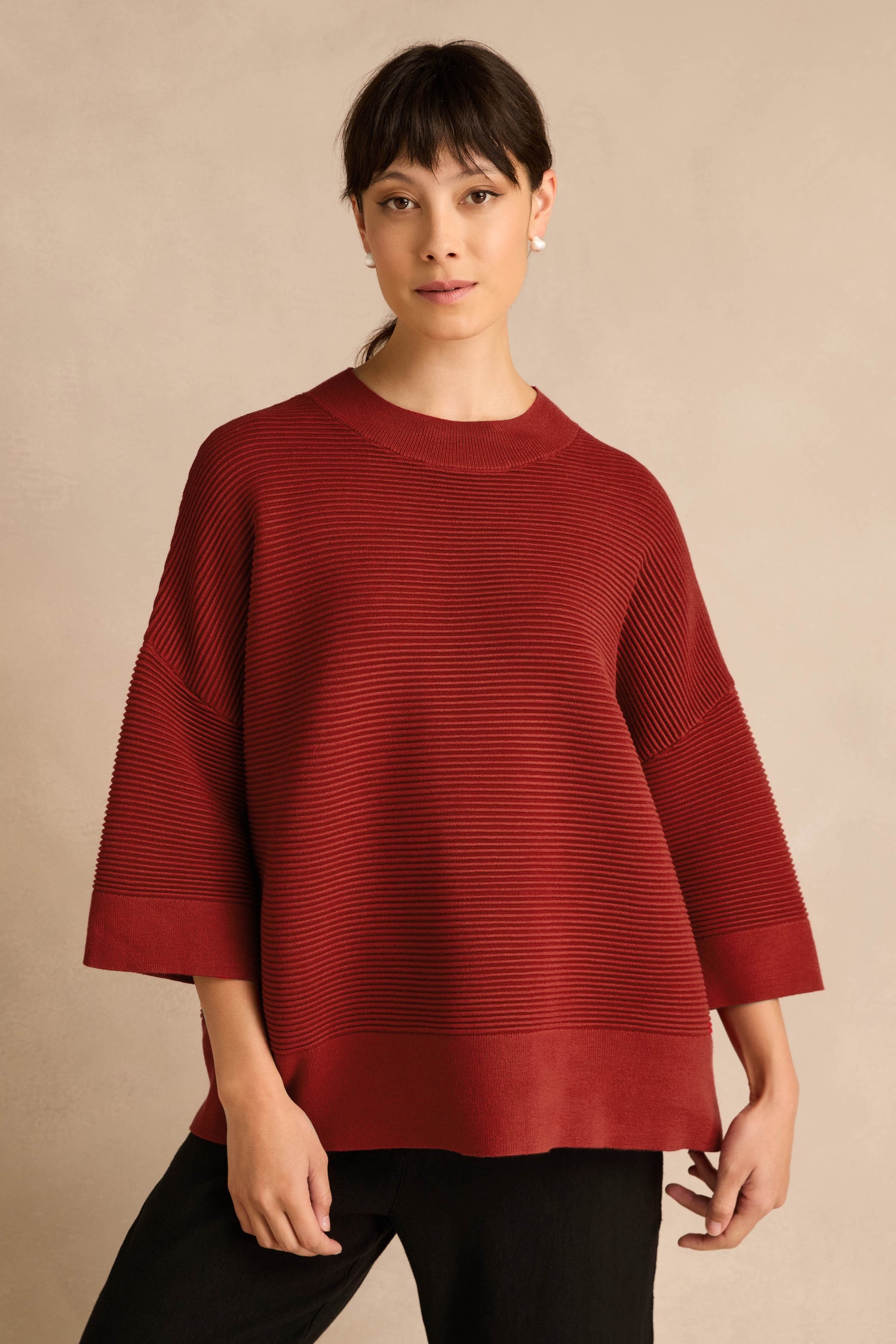 A person stands against a beige background wearing the Wide Sleeve Burgundy Ribbed Knit sweater paired with black pants.
