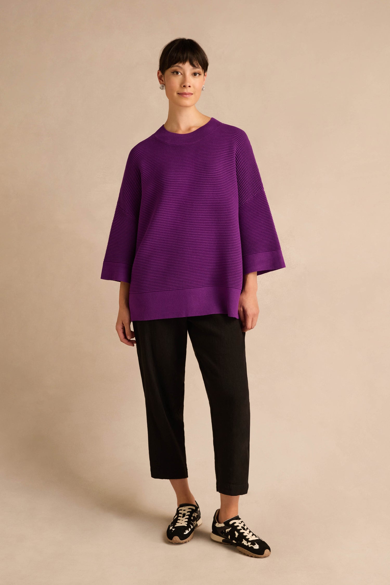 A person stands against a neutral background wearing the Wide Sleeve Purple Ribbed Knit, paired with black pants and black sneakers with white laces.