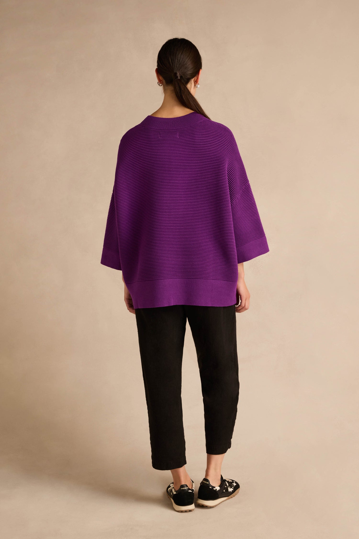 A person stands facing away against a plain beige background, wearing the Wide Sleeve Purple Ribbed Knit, paired with black pants and black shoes. Their long hair is tied back.