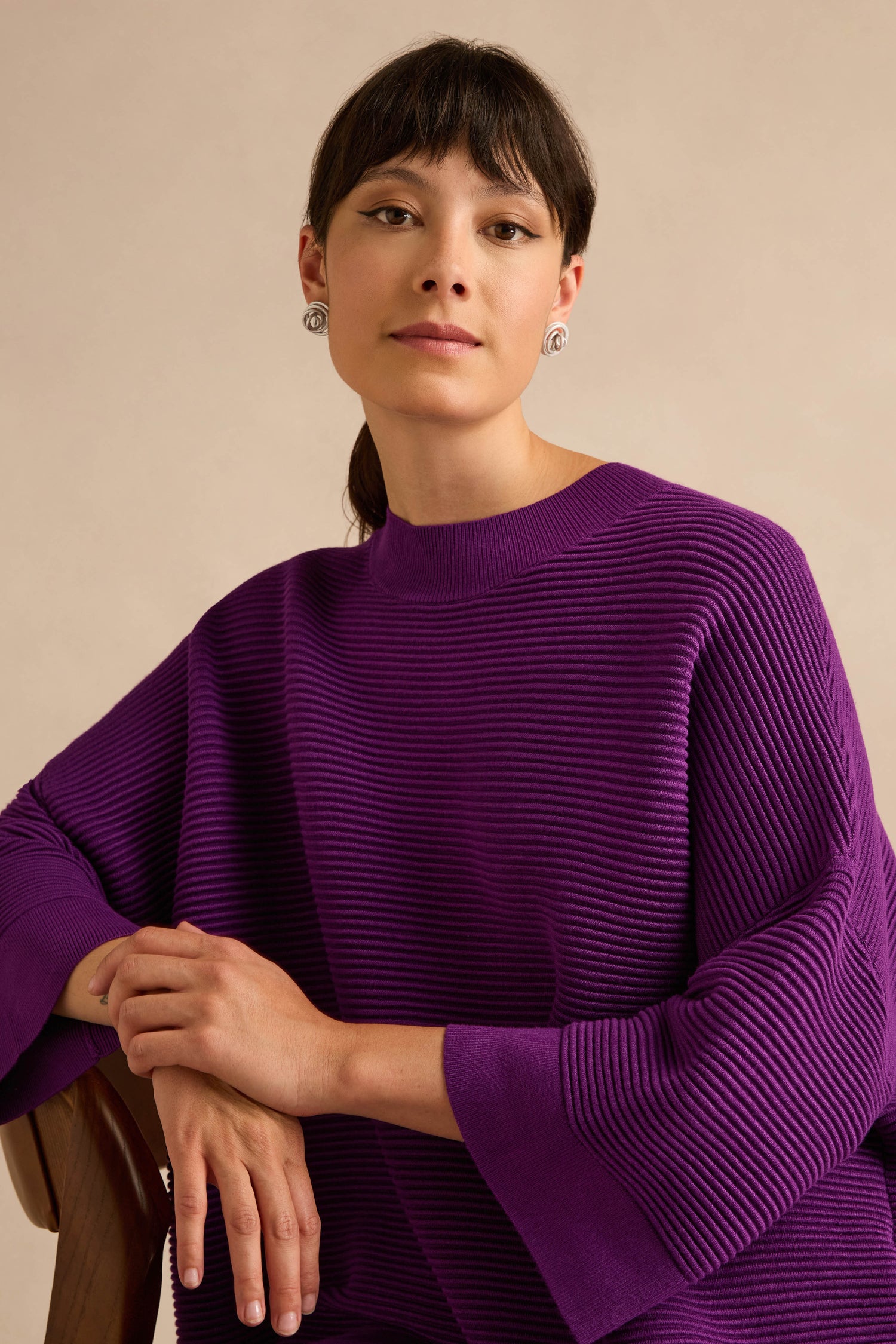 A person with dark hair and earrings wears the Wide Sleeve Purple Ribbed Knit in a relaxed fit, sitting with elbows resting on a chair against a neutral background.